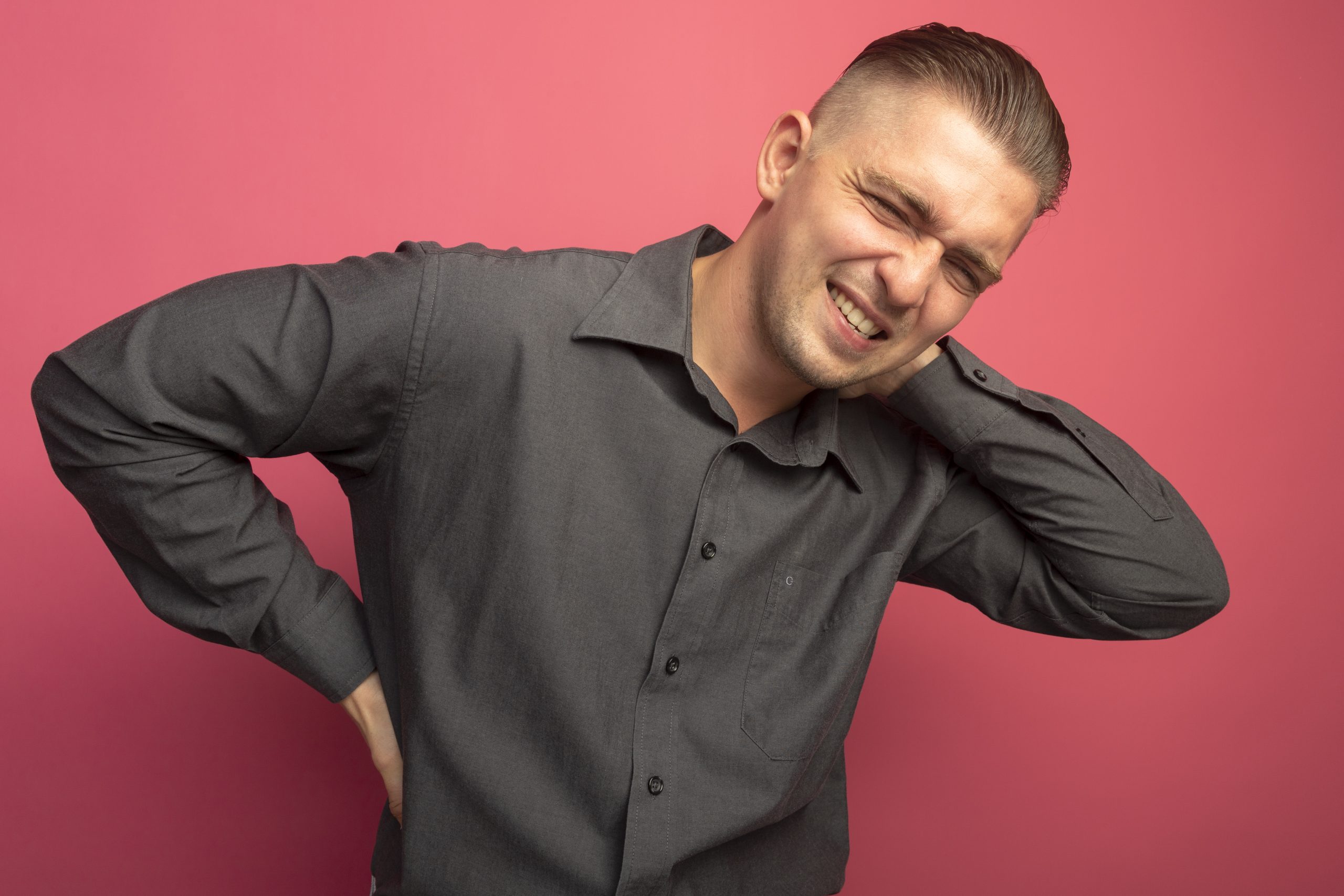young handsome man in grey shirt looking unwell touching his neck feeling pain standing over pink background